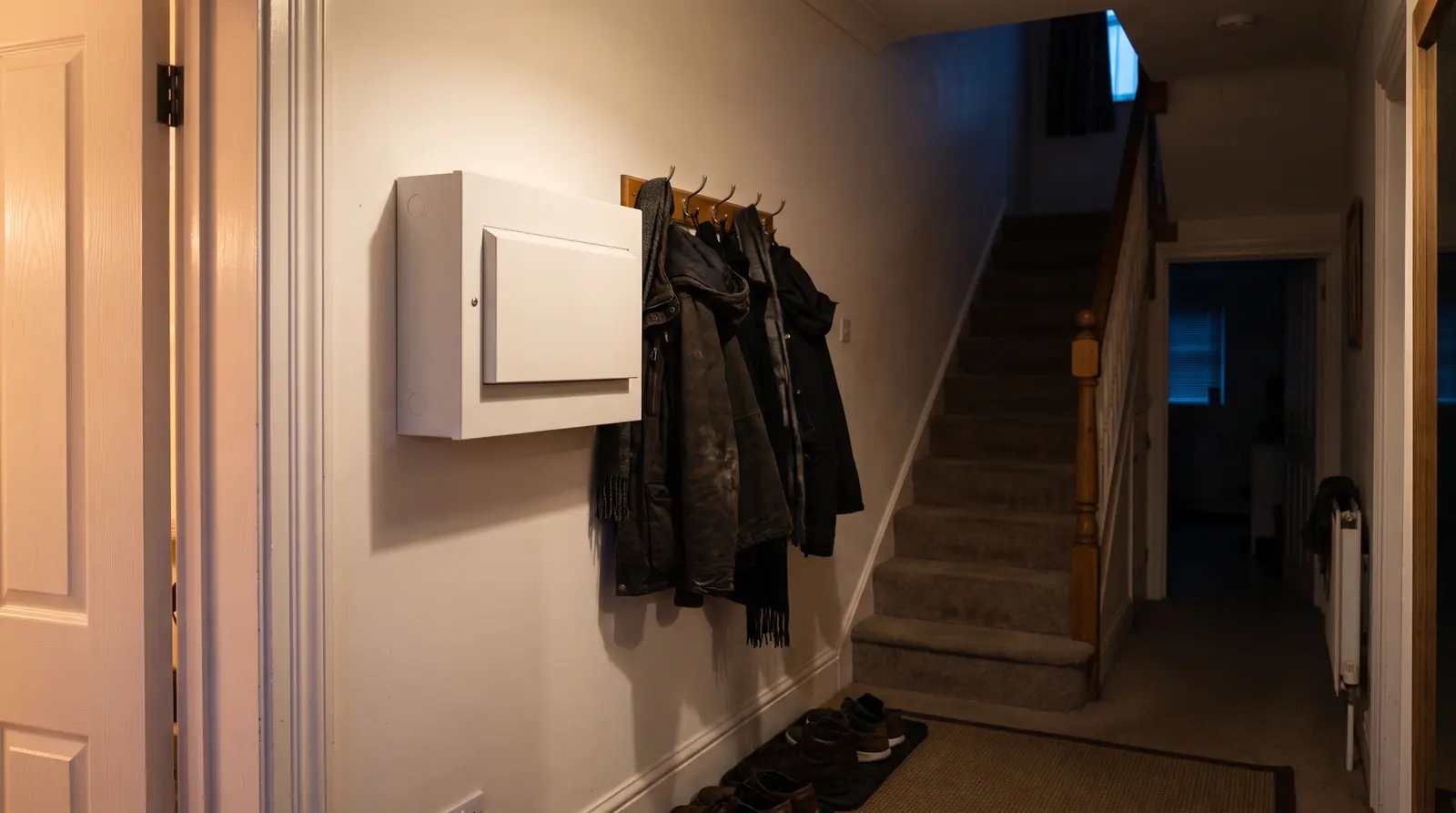 A modern consumer unit on the wall of a domestic hallway, coats on hooks and a staircase beyond