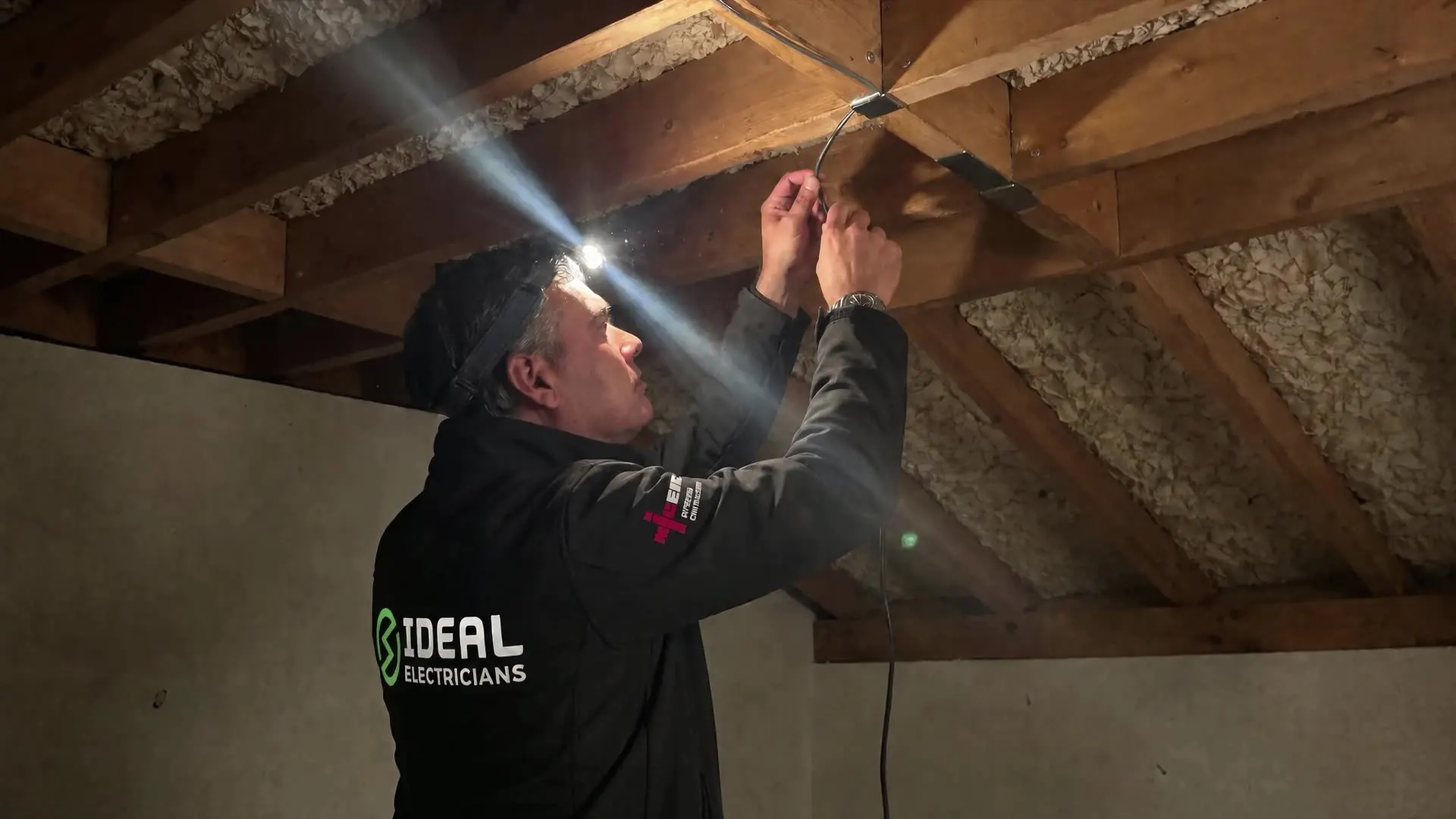 An Ideal Electricians engineer clipping cable to a rafter in a loft, lit by a head torch